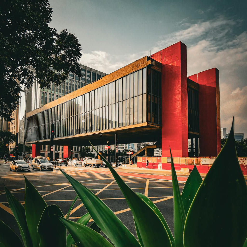 Foto/Ilustração do MASP (Museu de Arte de São Paulo), um edifício moderno com estrutura suspensa, fachada de vidro e pilares vermelhos, localizado na Avenida Paulista, com árvores, carros e céu parcialmente nublado ao fundo.