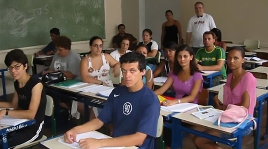 Grupo de estudantes em sala de aula, alguns com livros e cadernos, participando de aula escolar em um dia comum.