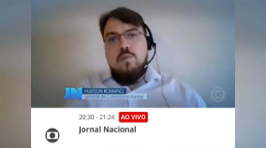 A man participating in a live television news interview, wearing glasses and headphones, with the Jornal Nacional logo highlighted in the background.