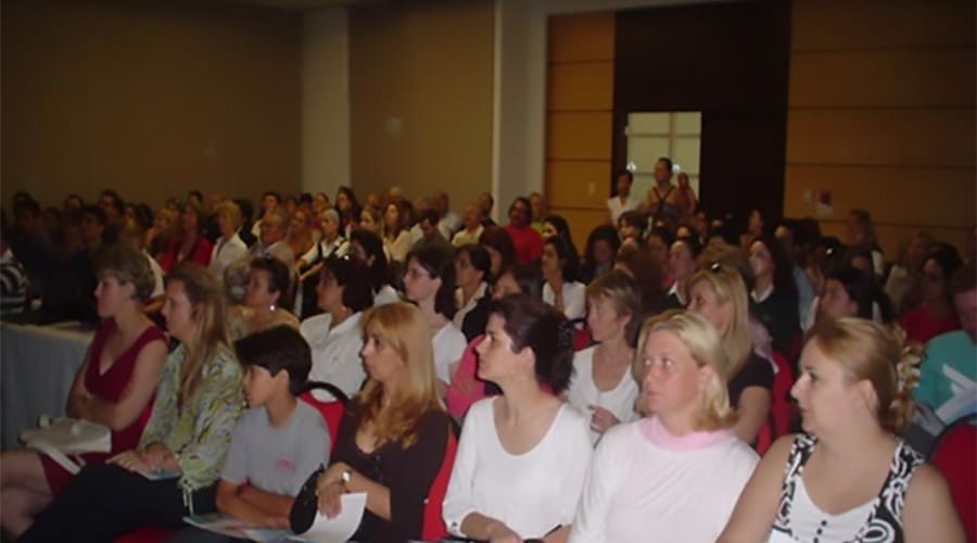 Grupo de pessoas assistindo a uma palestra ou evento em um auditório, várias idades e gêneros presentes, ambiente formal e bem iluminado.