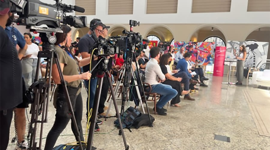 Pessoas assistindo a uma transmissão ao vivo de um evento, com câmeras profissionais e equipe de jornalistas em um ambiente de cobertura de mídia ao vivo.